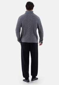 Grauer Strickpullover mit hohem Kragen, der eine gerippte Textur und ein zentrales Zopfdesign aufweist. Kombiniert mit lockeren schwarzen Hosen und dunklen Schuhen.