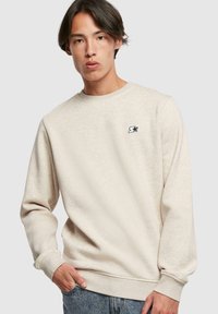 Sudadera beige con cuello redondo y puños acanalados, que presenta un pequeño logotipo de estrella negra en el pecho. Tejido suave con un acabado texturizado.