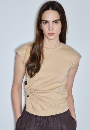 Jonge vrouw met lang bruin haar, gekleed in een beige mouwloos topje met plooien aan de zijkant en een donkere losse broek, staand tegen een effen lichte achtergrond.