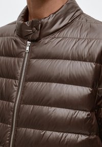 Doudoune marron avec un col montant, comprenant des panneaux cousus, une fermeture éclair frontale et une fermeture par boutons-pression. Texture lisse et brillante.