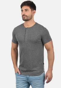 Grå kortærmet Henley skjorte lavet af en struktureret bomuldsblanding, med en tre-knaps knaplukning og en lige kant. Båret med lyseblå jeans.