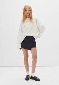 Přesně padnoucí bílý sweatshirt s oversize střihem a spadenými rameny, v kombinaci se přiléhavou černou sukní s proužky. Modelka má na sobě černé boty a bílé ponožky.