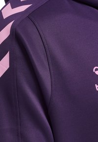 Giacca sportiva viola con dettagli a chevron rosa lungo la cucitura della spalla, tessuto testurizzato e finitura liscia. Logo visibile sul davanti.