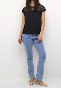 Donna vestita con una blusa nera in pizzo, jeans azzurri chiari e scarpe con tacco kitten beige, in piedi davanti a uno sfondo bianco.