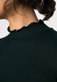 Mørkegrøn ribbet turtleneck med en tekstureret overflade og en flæset kant ved kraven. Nærbillede fremhæver halsområdet.