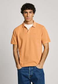 Oranžs polo krekls ar klasisku apkakli, īsām piedurknēm un diskrētu izšūtu logo uz krūšu daļas, kombinēts ar ziliem denima džinsiem.