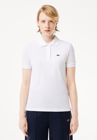 Weißes Polo-Shirt aus atmungsaktiver Stoff mit kurzen Ärmeln, versehen mit einem grünen, gestickten Logo auf der linken Brust und einem klassischen Kragen-Design.