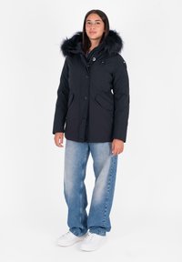 Zwarte winterjas met een gevoerde capuchon van bont, frontknopen en zijzakken. Bevat een getailleerd ontwerp met contrasterende texturen en materialen.