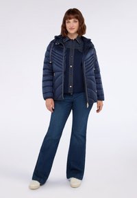 Donna in piedi che indossa un giubbotto trapuntato blu navy, una camicia in denim scuro, jeans a zampa d'elefante e sneaker bianche, su uno sfondo chiaro e semplice.