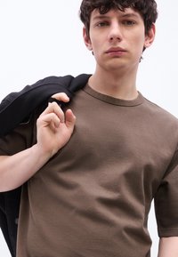 Νεαρός άνδρας με κοντά σγουρά μαλλιά, ντυμένος με καφέ μπλουζάκι και κρατώντας μία μαύρη ζακέτα στον ώμο του, μπροστά σε ένα απλό λευκό φόντο.