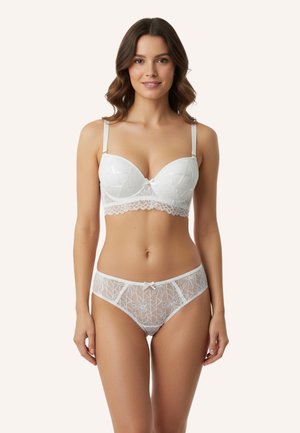 Kobieta w białym koronkowym komplecie bielizny, stojąca, zwrócona przodem, na jasnym, jednolitym tle, lekko się uśmiecha.