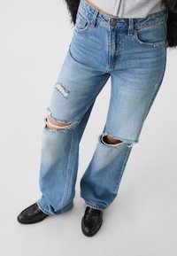 Siniset korkeavyötäröiset farkut leveillä lahkeilla, joissa kuluneita polvivaurioita ja rispaantuneet reunat, yhdistetty mustiin liukusovituskenkiin.