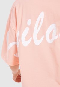 Ferskenfarvet bomulds t-shirt med løs pasform. Har stort hvidt kursiv tekst på ryggen, korte ærmer og en blød tekstur.