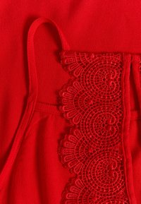 Červený top z látky s propracovaným krajkovým detailem podél výstřihu, tenkými ramínky a hladkou strukturou po celé ploše.