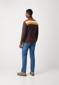 Brauner Pullover mit hohem Kragen, der auffällige horizontale Streifen in Orange, Weiß und Gelb über den Schultern aufweist. Kombiniert mit blauen Jeans.