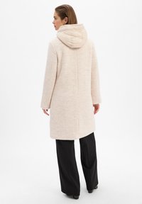 Lichtbeige textuur jas met een gewatteerde capuchon, rechte silhouet, lange mouwen en zijzakken. Gepaard met zwarte wijde broek.