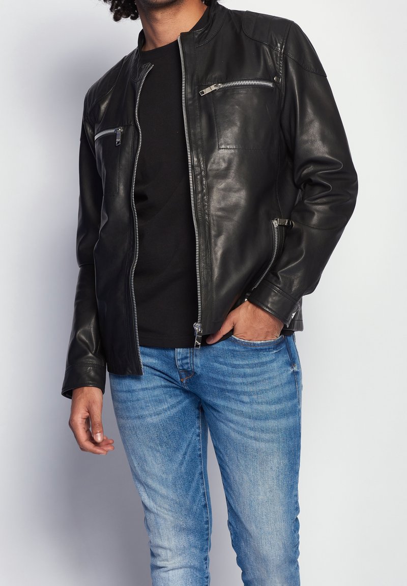 Giacca di pelle nera con colletto alto, chiusura con zip e due tasche sul petto con zip. Abbinata a jeans blu con una vestibilità slim.