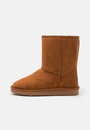 Botte d'hiver mi-mollet en daim brun avec semelle plate rainurée et coutures apparentes, vue de côté extérieur sur fond blanc.