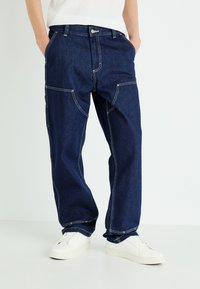 Pantaloni in denim blu scuro con vestibilità ampia, caratterizzati da cuciture bianche, tasche frontali e tasche cargo laterali. Abbinati a sneakers bianche.