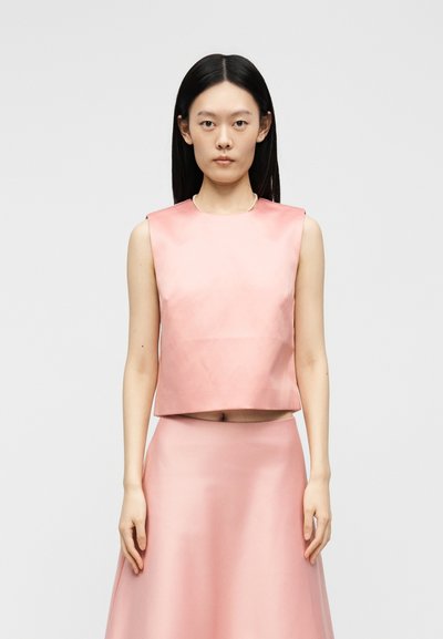 Γυναίκα με μακριά μαύρα μαλλιά που φοράει ένα ροζ αμάνικο σατέν crop top και μια ταιριαστή ψηλόμεση φούστα, στέκεται μπροστά σε ένα απλό φωτεινό φόντο.