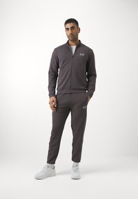 Completo tuta grigio scuro con giacca con zip e pantaloni affusolati abbinati. Presenta il logo EA7. Indossato con sneaker bianche. Texture liscia.