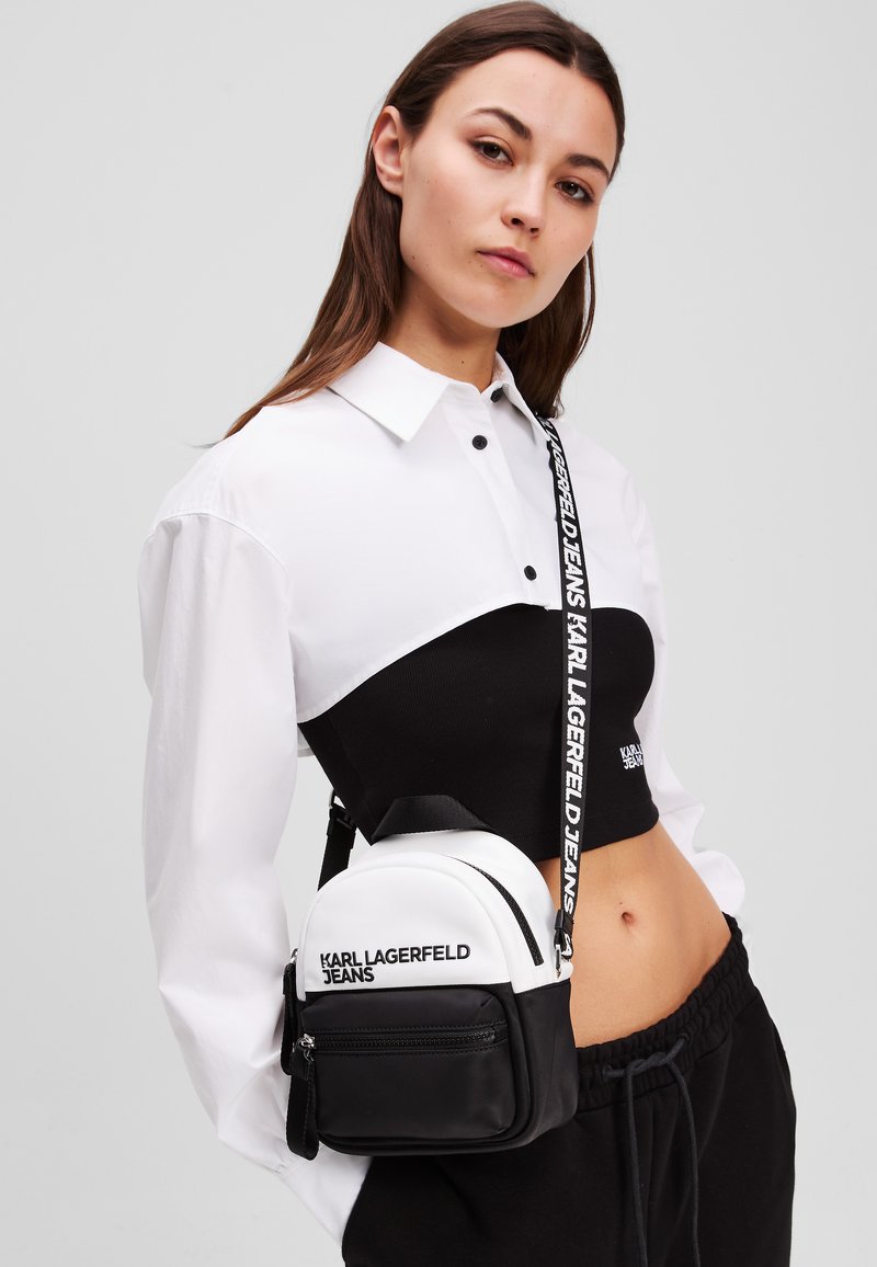 Zaino mini realizzato in tessuto bianco e nero, con una forma arrotondata, pannelli a contrasto, una tracolla con logo e una tasca frontale con zip.