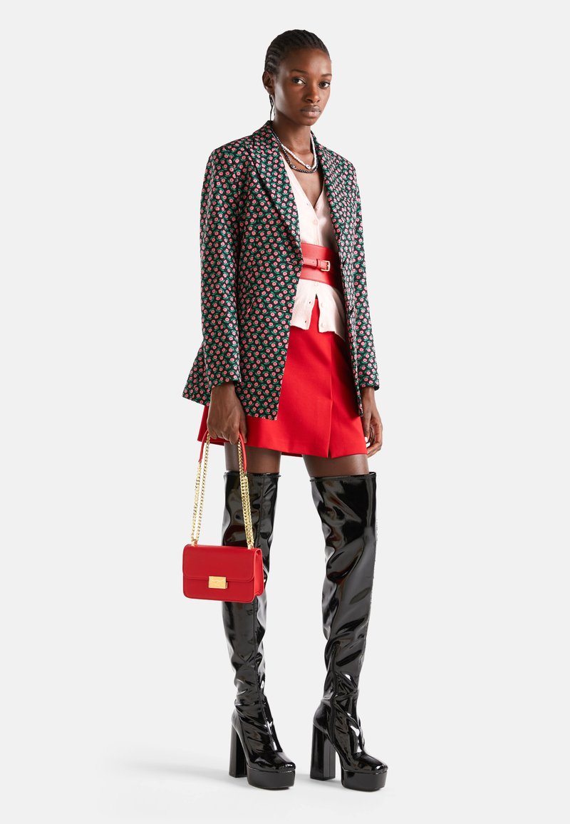 Blazer à motifs multicolores, blouse rose, jupe rouge, bottes noires montantes et sac à main rouge avec une bandoulière en chaîne. Textures lisses tout au long.