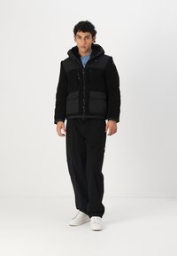 Sort jakke med en tekstureret fleece-krop og ensartede stofærmer, med lynlås, lommer og hætte. Sammen med sorte bukser og hvide sko.