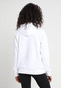 Una persona con capelli ricci si trova di spalle indossando una felpa bianca semplice e leggings neri su uno sfondo chiaro.