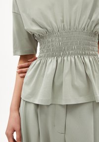 Světle zelený top s nabíraným pasem a krátkými rukávy, s rozšířeným lemem. Látka vypadá měkká s hladkým povrchem.