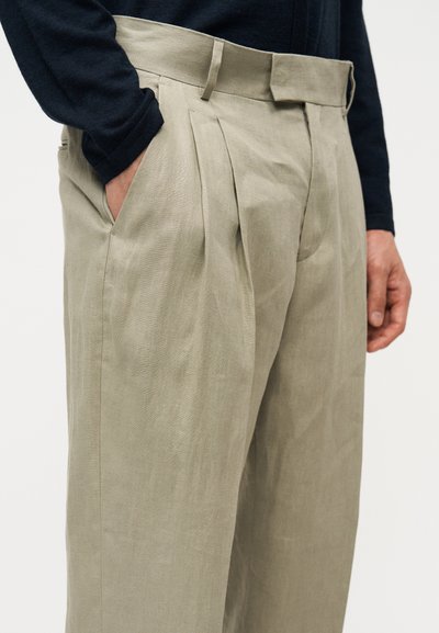 Μπεζ πλισέ παντελόνια φορεμένα με μαύρη μπλούζα μακρυμάνικη, με το ένα χέρι στην τσέπη, εμφανιζόμενα από το μέσο του μηρού έως τη μέση.