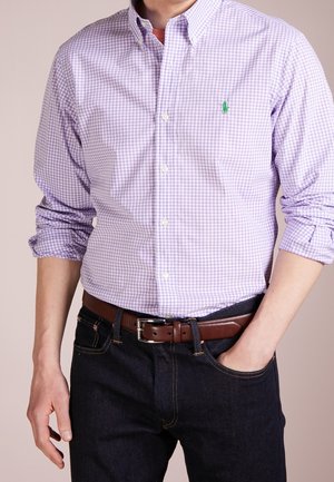 Mies, joka pukeutunut laventelin- ja valkoraidalliseen ruutupaitaan, jossa vihreä logo, tummiin farkkuihin, ruskeaan nahkavyöhön, käsi taskussa.