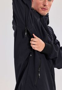 Czarna kurtka typu softshell z zamkami błyskawicznymi po bokach i na rękawach. Posiada wysoki kołnierz i ściągacze na mankietach. Gładki materiał z minimalną liczbą szwów.