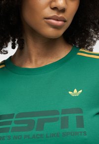 Κοντινό πλάνο ενός ατόμου που φοράει πράσινη αθλητική μπλούζα Adidas με κίτρινες ρίγες και αθλητικό λογότυπο κοντά στον γιακά.