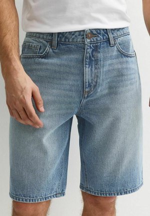 Hombre con pantalones cortos de mezclilla azul claro y camiseta blanca, de pie con los brazos relajados a los lados contra un fondo liso.