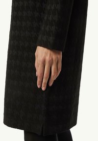 Schwarzer, strukturierter Mantel mit einem geometrischen Muster, der eine Nahaufnahme des Ärmels und der Hand zeigt, die auf dem Stoff positioniert ist.