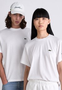 Dva modelové v bílých tričkách Lacoste s logem krokodýla; muž s kudrnatými vlasy a čepicí, žena s dlouhými rovnými černými vlasy, neutrální výrazy.