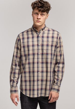 Młody mężczyzna z kręconymi włosami, ubrany w beżową, niebieską i czerwoną koszulę w kratę z guzikami oraz ciemne dżinsy, na jednolitym tle.