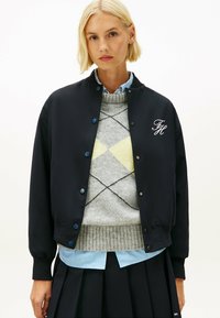 Marine bomberjack met blauwe drukknoopjes, geborduurde initialen en geribbelde manchetten over een grijs ruitjestrui en gestreept shirt.