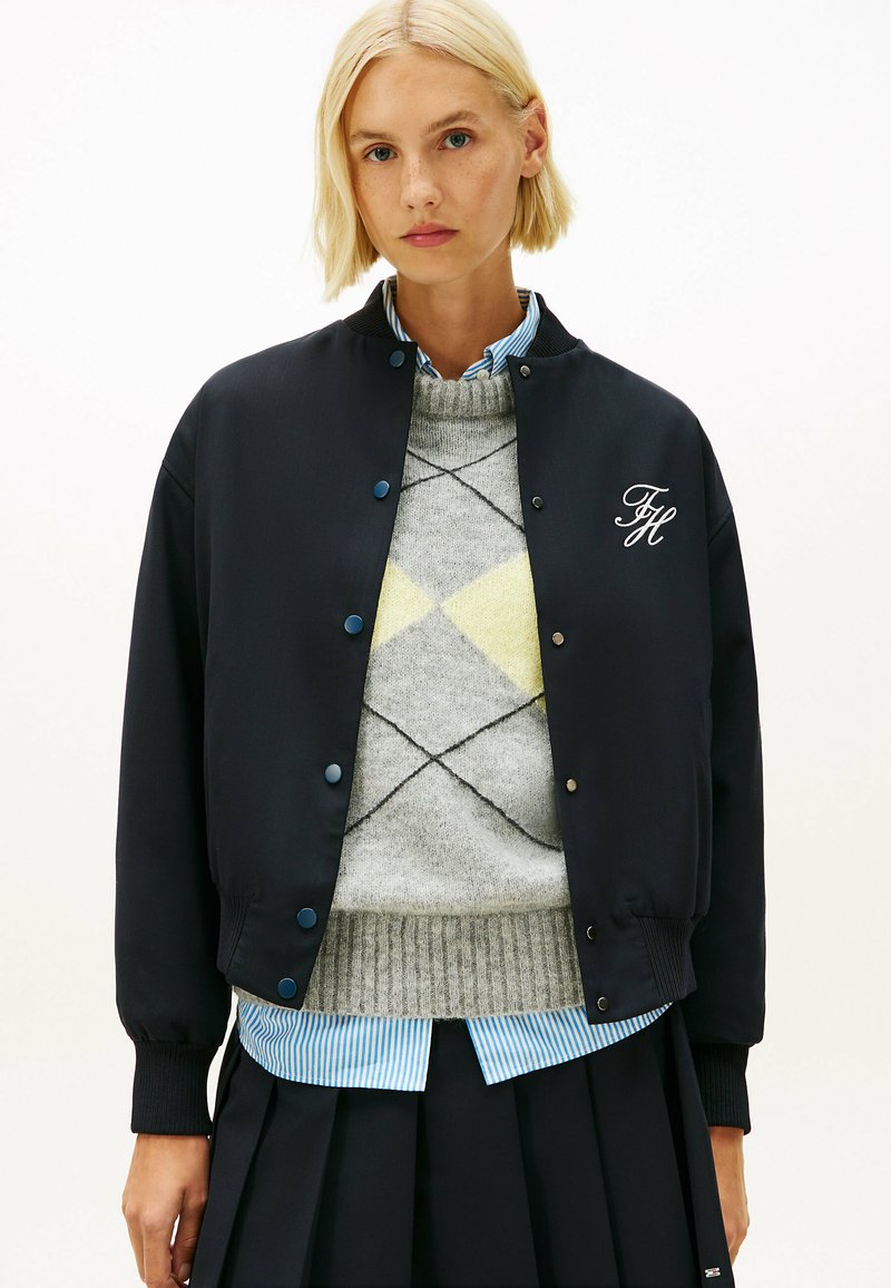 Marine bomberjack met blauwe drukknoopjes, geborduurde initialen en geribbelde manchetten over een grijs ruitjestrui en gestreept shirt.