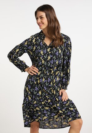 Vrouw glimlacht en poseert in een lange mouw zwarte jurk met geel en paars bloemenpatroon tegen een effen witte achtergrond.
