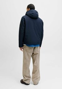Veste à capuche navy au design épuré, associée à un pantalon large beige clair et des chaussures sombres, vue de l'arrière.