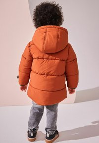 Doudoune orange avec capuche, présentant un design matelassé, portée avec un pantalon gris. Des baskets noires et blanches avec semelles en caoutchouc sont visibles.