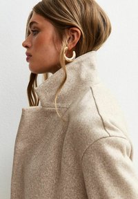 Manteau en mélange de laine beige avec un col haut et un design minimaliste. Présente un tissu texturé et des détails de couture subtils.