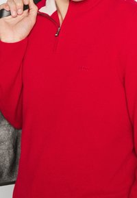 Pull rouge à fermeture éclair fabriqué en matière tricotée douce, avec un col en polaire blanche contrastante et un logo embossé subtil sur la poitrine.