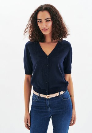 Cardigan marine à col en V, à manches courtes, avec un détail texturé floral, associé à une ceinture de couleur claire et des jeans en denim.