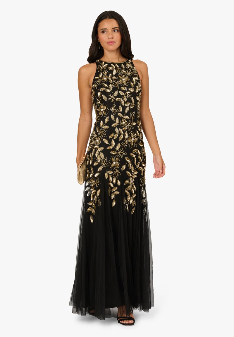 Žena v dlouhých černých šatech bez rukávů se zlatou květinovou výšivkou, držící zlatou kabelku a černé sandály na vysokém podpatku.