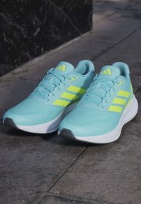 Baskets de sport bleu clair avec trois bandes jaunes, dessus en maille texturée, col rembourré et semelle en caoutchouc avec des accents gris.