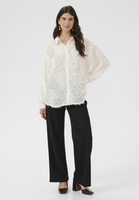 Prozorna bela bluza z reliefnim cvetličnim vzorcem, gumbe na sprednji strani in dolgimi rokavi, v kombinaciji s črnimi širokimi hlačami in bordo petami.