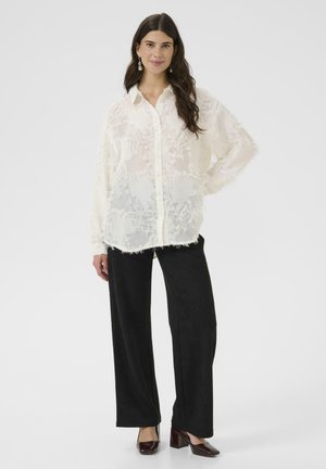Prosojna bele bluza z teksturnim cvetličnim vzorcem, z gumbom spredaj, sproščen kroj, dolgi rokavi, v kombinaciji z črnimi širokimi hlačami in temnimi čevlji.